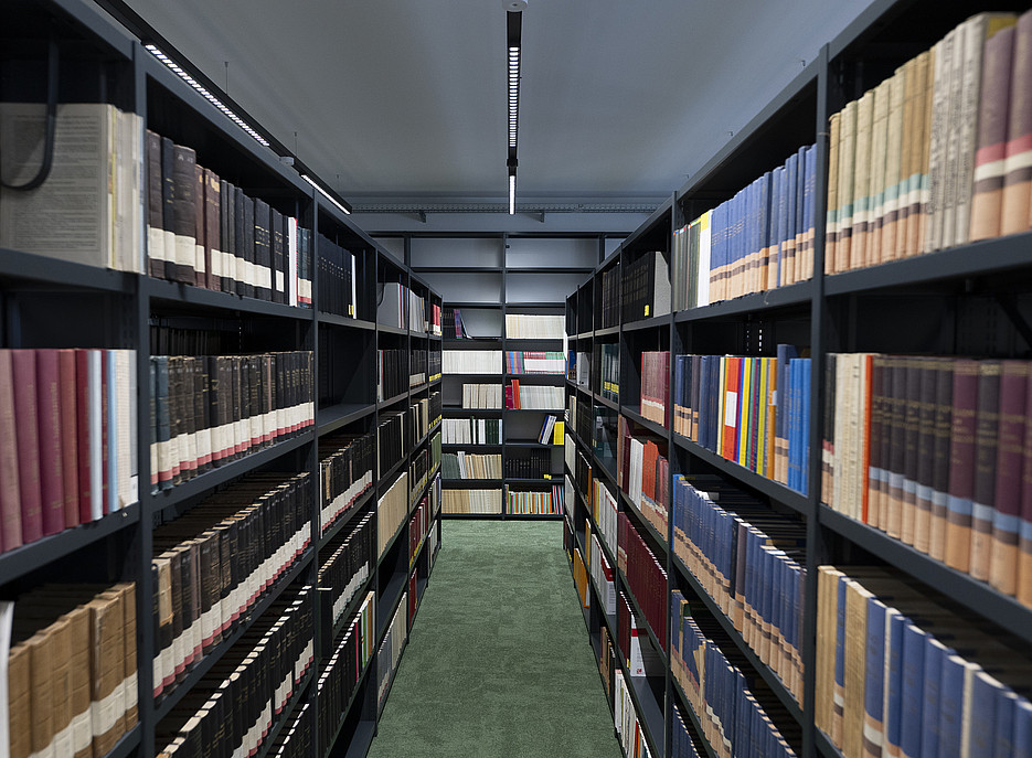 „Gang zwischen hohen, dunkelgrauen Bücherregalen in einer Bibliothek, dicht gefüllt mit Büchern in unterschiedlichen Farben; über dem grünen Teppichboden verläuft mittig eine schmale LED-Deckenleuchte. ©Uni Graz/Tzivanopoulos