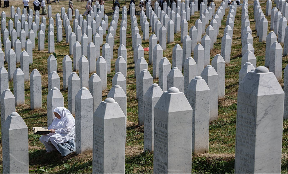 Eine Frau in weißer Kleidung sitzt zwischen Reihen weißer Grabsteine auf dem Friedhof der Gedenkstätte Srebrenica-Potočari und liest in einem Buch.