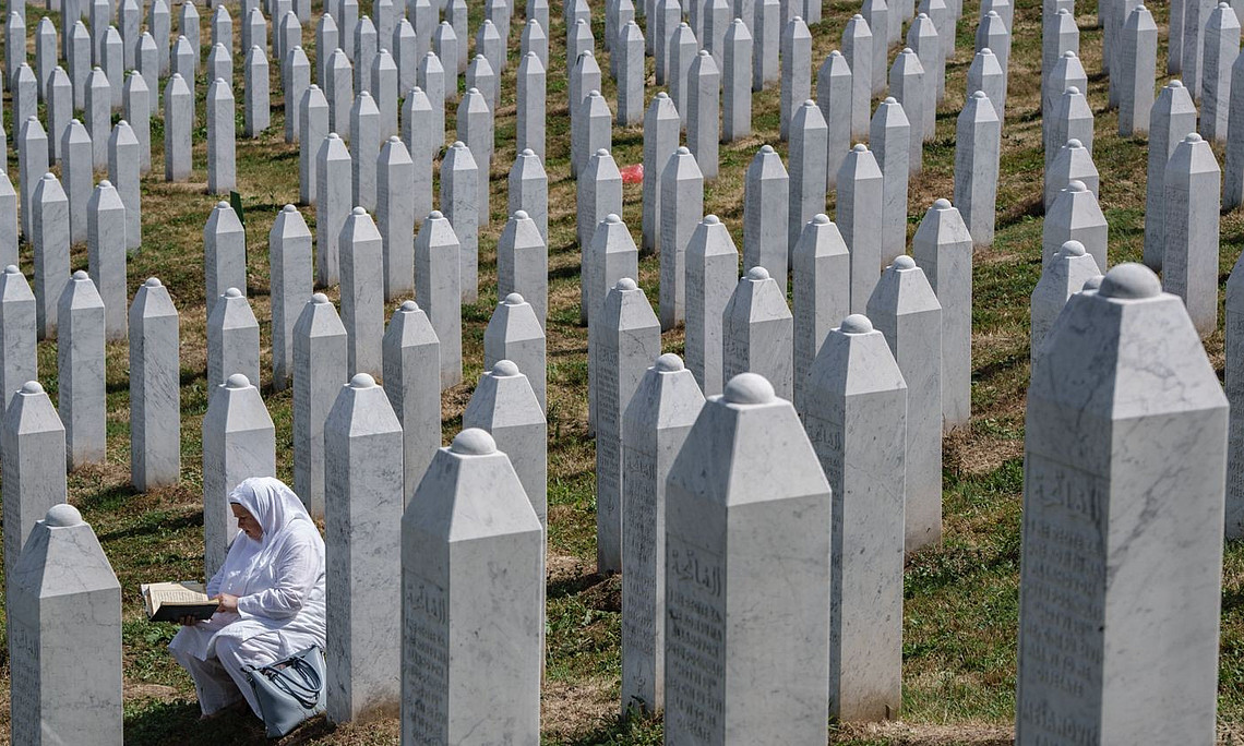 Eine Frau in weißer Kleidung sitzt zwischen Reihen weißer Grabsteine auf dem Friedhof der Gedenkstätte Srebrenica-Potočari und liest in einem Buch.