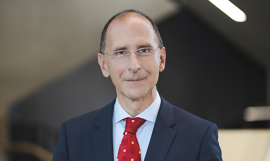 A middle-aged man is standing in front of a flight of stairs in a modern building. He is wearing a dark blue suit, a light blue shirt and a red patterned tie. The man has short hair, wears glasses and has his hands clasped in front of his body. The atmosphere appears professional and serious. ©Uni Graz/Tzivanopoulos