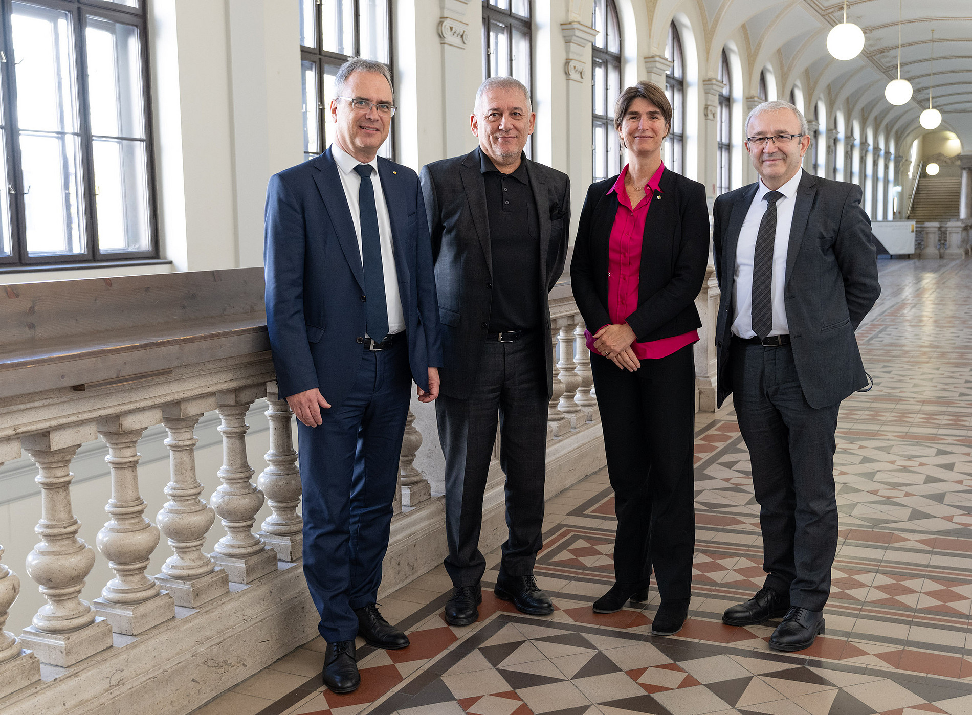Vier Personen stehen nebeneinander in einem historischen Gebäude mit hohen Decken, großen Fenstern und einem gemusterten Fliesenboden. Im Hintergrund sind eine Balustrade und ein langer Gang mit Rundbögen und hängenden Lampen zu sehen. Die Personen tragen formelle Kleidung, darunter Anzüge und Blazer. 
