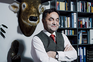 A person in formal attire with their arms crossed stands in front of a bookshelf, an artificial animal head hangs on the wall, and a cactus in a pot stands on a cabinet.