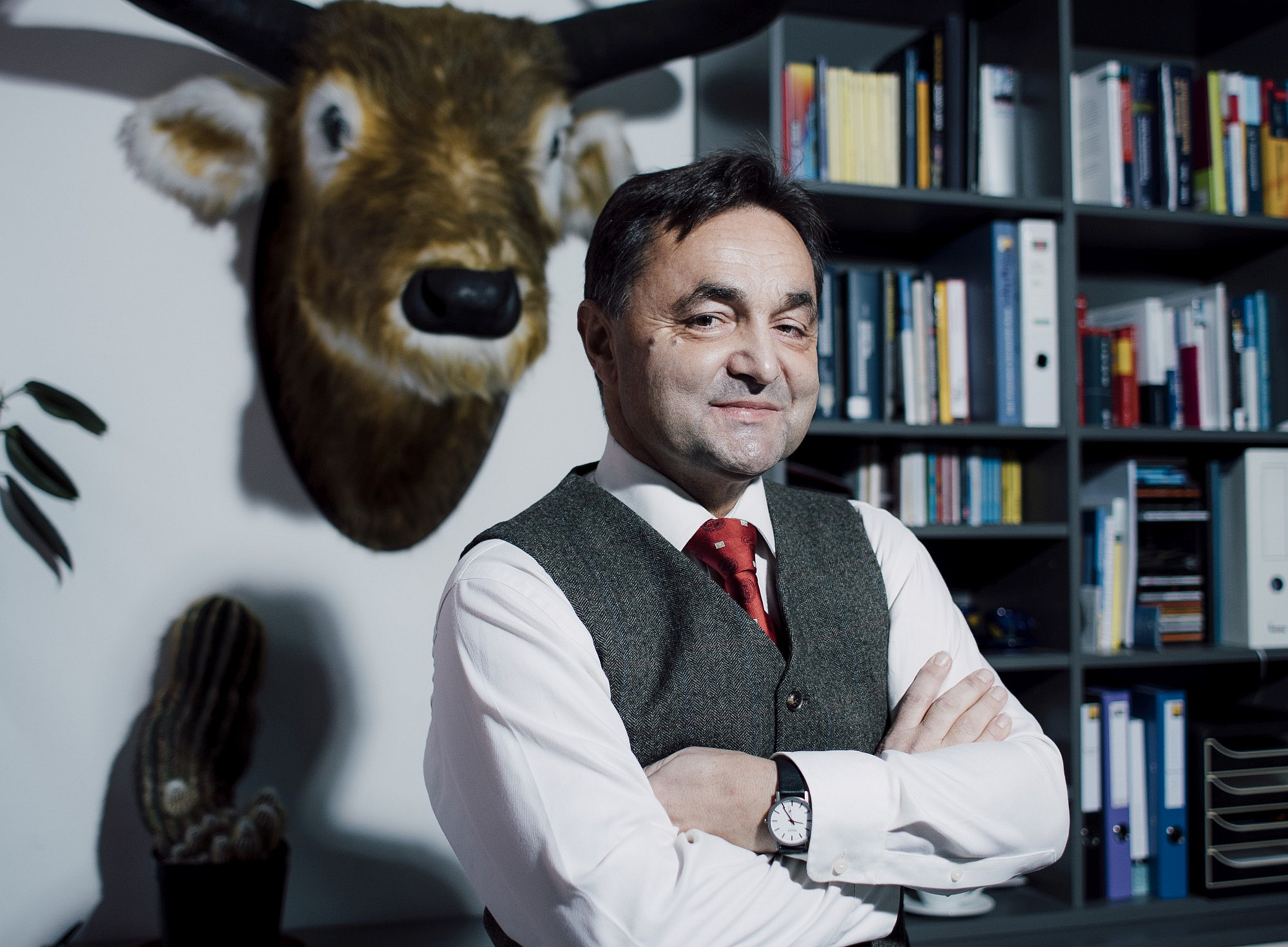 Eine Person in formeller Kleidung mit verschränkten Armen steht vor einem Bücherregal, an der Wand hängt ein künstlicher Tierkopf, und auf einem Schrank steht ein Kaktus in einem Topf. 