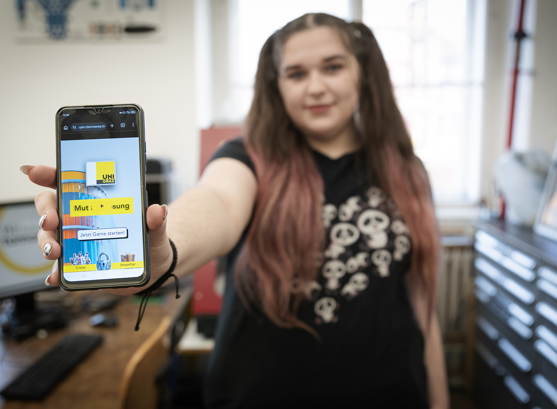 Person steht in einem Büro oder Arbeitsraum und streckt einen Arm nach vorne, um ein Smartphone in die Kamera zu halten. Auf dem Smartphone ist eine bunte Benutzeroberfläche mit dem gelben Logo „UNI GRAZ“, dem Schriftzug „Mut : Lösung“, einem Button „Jetzt Game starten!“ zu sehen. Die Person trägt ein dunkles T-Shirt mit mehreren stilisierten Pandabär-Motiven, im Hintergrund sind ein Schreibtisch mit Computer, Regale und weitere Büroeinrichtung erkennbar. ©Uni Graz/Angele