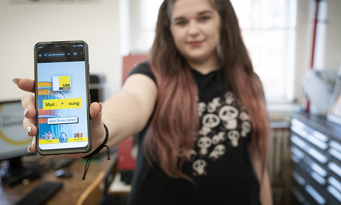 Person steht in einem Büro oder Arbeitsraum und streckt einen Arm nach vorne, um ein Smartphone in die Kamera zu halten. Auf dem Smartphone ist eine bunte Benutzeroberfläche mit dem gelben Logo „UNI GRAZ“, dem Schriftzug „Mut : Lösung“, einem Button „Jetzt Game starten!“ zu sehen. Die Person trägt ein dunkles T-Shirt mit mehreren stilisierten Pandabär-Motiven, im Hintergrund sind ein Schreibtisch mit Computer, Regale und weitere Büroeinrichtung erkennbar.