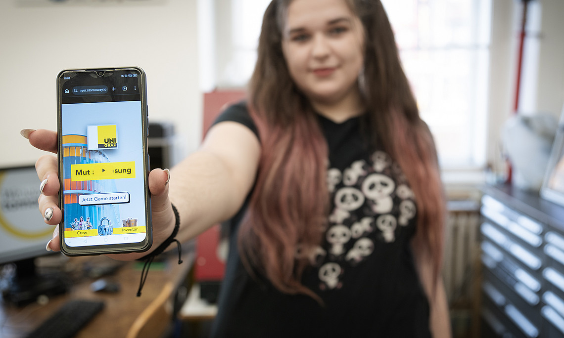 A person stands in an office or workroom and stretches one arm forward to hold a smartphone up to the camera. The smartphone displays a colourful user interface with the yellow ‘UNI GRAZ’ logo, the words ‘Courage: Solution’ and a button labelled ‘Start game now!’. The person is wearing a dark T-shirt with several stylised panda bear motifs. In the background, a desk with a computer, shelves and other office furniture can be seen.