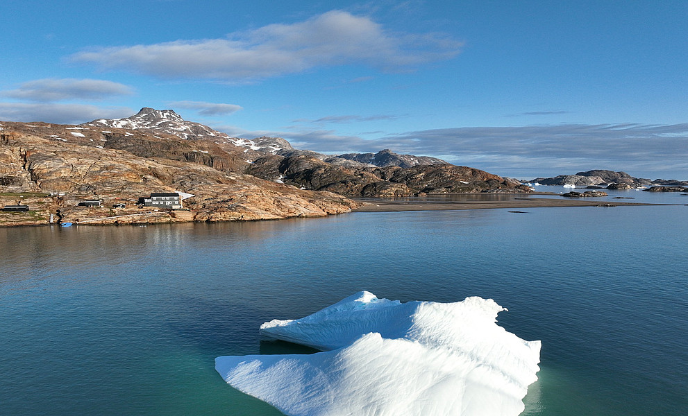 Ein Eisberg im Vordergrund, der türkisblaues Schmelzwasser umspült wird; im Hintergrund steile, teilweise schneebedeckte Felsberge, eine kleine Forschungsstation am Ufer und ein langer Holzsteg, der ins Meer ragt – klare, kühle Farben bei wolkenlosem Himmel, aufgenommen aus der Vogelperspektive.