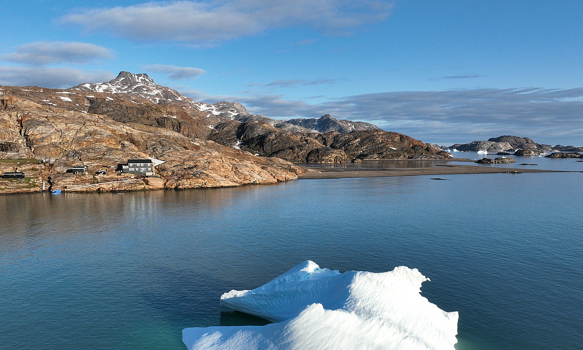 Ein Eisberg im Vordergrund, der türkisblaues Schmelzwasser umspült wird; im Hintergrund steile, teilweise schneebedeckte Felsberge, eine kleine Forschungsstation am Ufer und ein langer Holzsteg, der ins Meer ragt – klare, kühle Farben bei wolkenlosem Himmel, aufgenommen aus der Vogelperspektive.