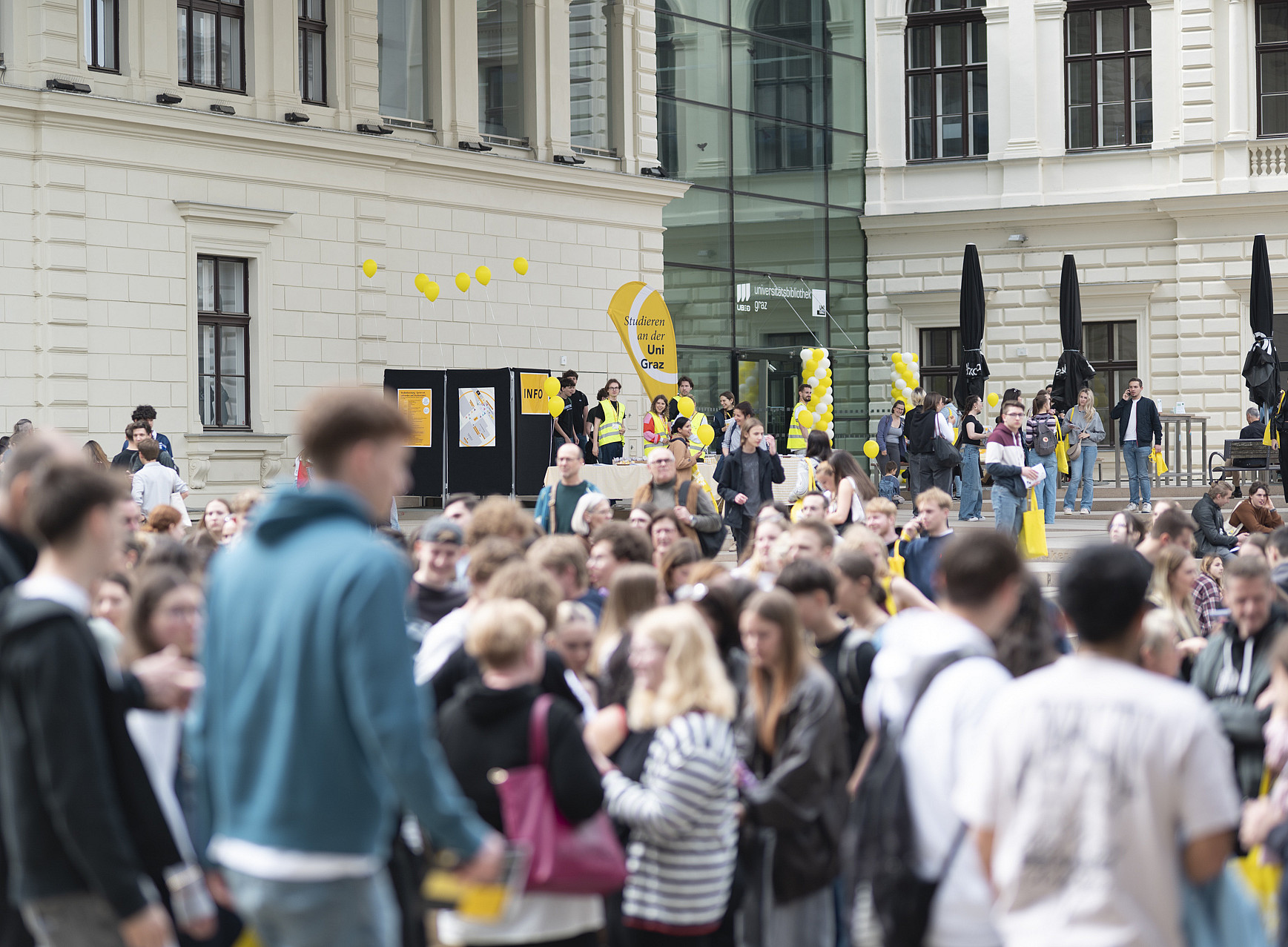 A large crowd stands and walks in a square in front of a multi-storey, bright building with tall windows; yellow balloons, information stands with yellow signs and a glass façade can be seen in the background. ©Uni Graz/Angele