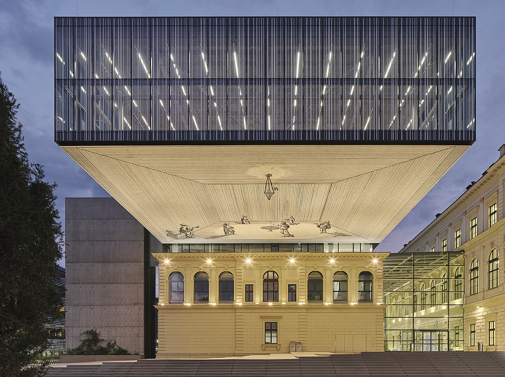 Exterior view of the University Library of the University of Graz at night, brightly lit exterior façade and the ceiling construction with a large mural painting ©Schreyer David