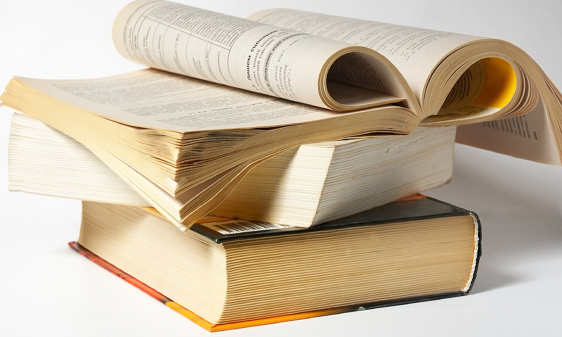 Drei Bücher liegen übereinander. Das oberste Buch ist aufgeschlagen und die Seiten sind eingerollt. 