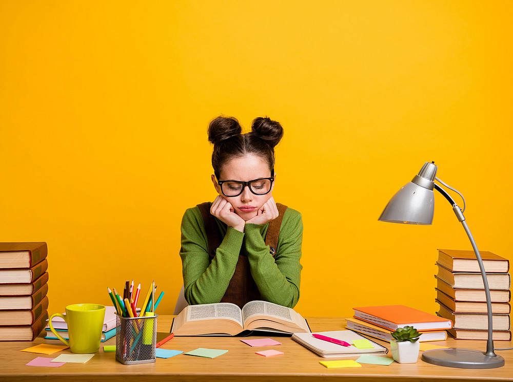 Junge Frau mit Zöpfen sitzt konzentriert an einem Schreibtisch vor gelbem Hintergrund und symbolisiert die Entwicklung wissenschaftlicher Textkompetenz. ©deagreez - stock.adobe.com 