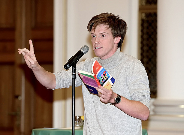 Christoph Steiner in front of the microphone, a book in his hand, his right index finger raised ©CP-PICTURES