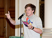 Christoph Steiner in front of the microphone, a book in his hand, his right index finger raised