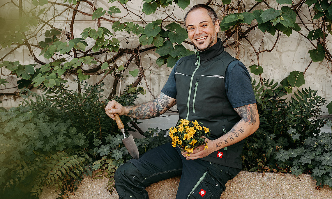 Gärtner Thomas Galik sitzt mit Schaufel und Pflanze in der Hand auf einem bepflanzten Trog
