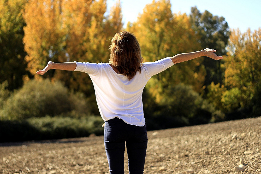 Eine Person steht in der Natur auf einer Wiese, die Arme weit von sich gestreckt. Im Hintergrund sind Bäume und Büsche. Der Himmel ist blau. ©pixabay/alfcermed