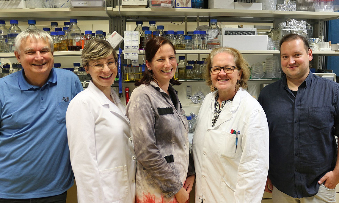5 scientists standing in front of the laboratory, one man with a blue shirt, one woman with short blonde hair wearing a labor cote, one woman with long brown hair and a grey blouse, one women wearing Labor cote , on man has short dark hair dressed in navy-blue shirt jeans, laboratory full of shelves filled up by test tubes and small plastic containers with different glowing colors inside