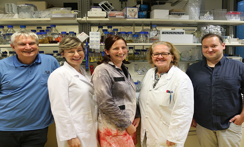 5 Wissenschaftler stehen vor dem Labor, ein Mann mit einem blauen Hemd, eine Frau mit kurzen blonden Haaren und einem Laborkittel, eine Frau mit langen braunen Haaren und einer grauen Bluse, eine Frau mit Laborkittel, ein Mann mit kurzen dunklen Haaren, einem dunkelblauen Hemd und Jeans, ein Labor voller Regale, die mit Reagenzgläsern und kleinen Plastikbehältern in verschiedenen leuchtenden Farben gefüllt sind