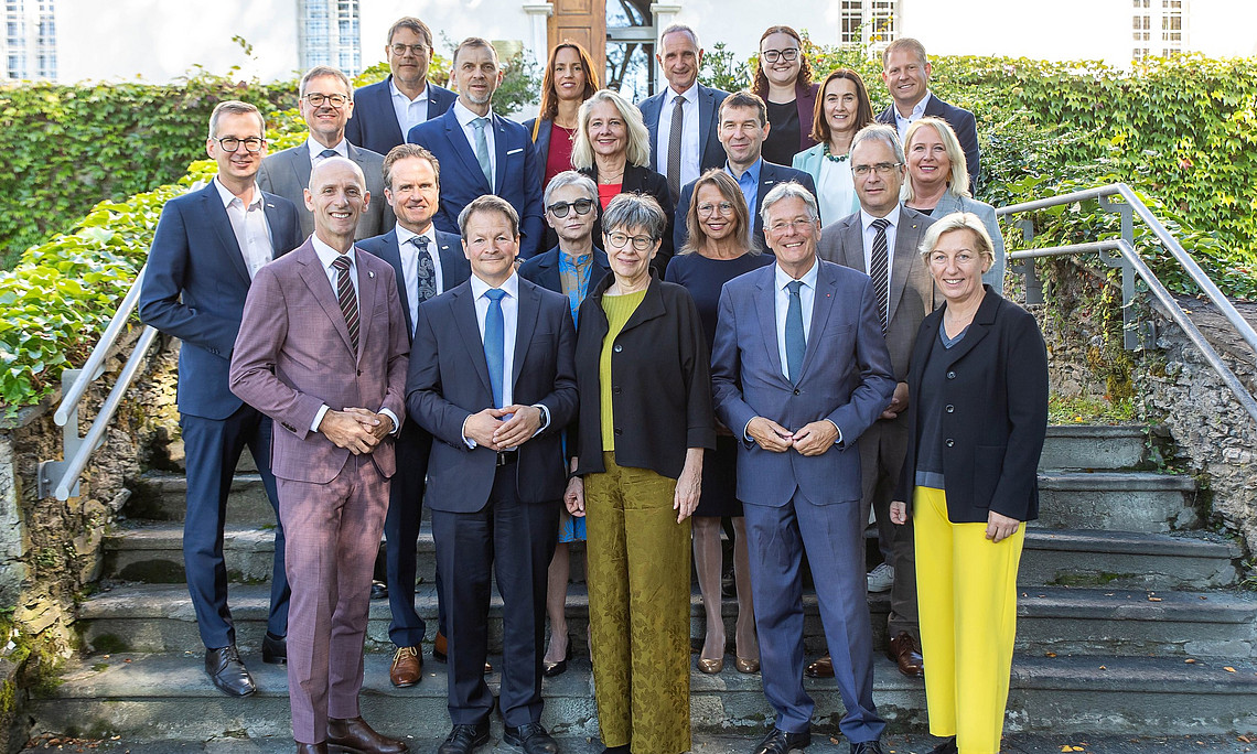 Ein Gruppenfoto der Politiker und Universitätsvertreter:innen bei einem Treffen in Klagenfurt, die Anzüge mit gelben oder blauen Akzenten tragen und auf Steinstufen vor einem alten weißen Gebäude stehen, das von Grün umgeben ist. Die Männer tragen dunkle Anzugjacken und Krawatten, während die Frauen lange Kleider in verschiedenen Farben tragen, darunter Schwarz, Grau, Rosa, Rot, Marineblau, Lila, Hellbraun, Gelb und Weiß. Sie posieren für Fotos, lächeln und schauen direkt in die Kamera.