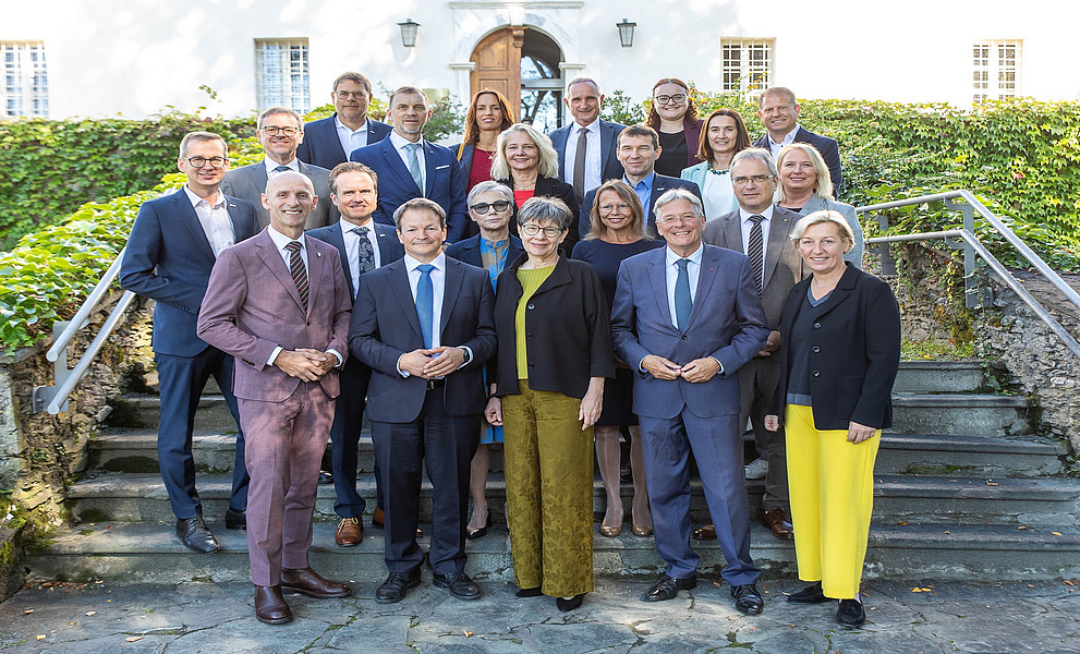 Ein Gruppenfoto der Politiker und Universitätsvertreter:innen bei einem Treffen in Klagenfurt, die Anzüge mit gelben oder blauen Akzenten tragen und auf Steinstufen vor einem alten weißen Gebäude stehen, das von Grün umgeben ist. Die Männer tragen dunkle Anzugjacken und Krawatten, während die Frauen lange Kleider in verschiedenen Farben tragen, darunter Schwarz, Grau, Rosa, Rot, Marineblau, Lila, Hellbraun, Gelb und Weiß. Sie posieren für Fotos, lächeln und schauen direkt in die Kamera.