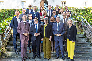 A group photo of politicians and university representatives at a meeting in Klagenfurt, wearing suits with yellow or blue accents and standing on stone steps in front of an old white building surrounded by greenery. The men are wearing dark suit jackets and ties, while the women are wearing long dresses in various colors, including black, gray, pink, red, navy blue, purple, light brown, yellow, and white. They are posing for photos, smiling and looking directly at the camera.