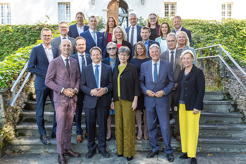 A group photo of politicians and university representatives at a meeting in Klagenfurt, wearing suits with yellow or blue accents and standing on stone steps in front of an old white building surrounded by greenery. The men are wearing dark suit jackets and ties, while the women are wearing long dresses in various colors, including black, gray, pink, red, navy blue, purple, light brown, yellow, and white. They are posing for photos, smiling and looking directly at the camera.