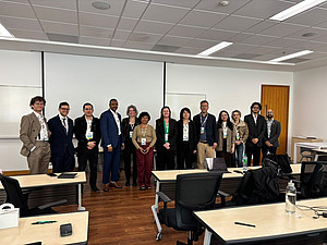 Gruppenfoto von zwölf Teilnehmenden einer internationalen Fachtagung. Die Gruppe steht nebeneinander in einem Seminarraum vor einer weißen Wand. Im Vordergrund sind Tische und Stühle des Seminarraums zu sehen. ©Paul Schweighofer