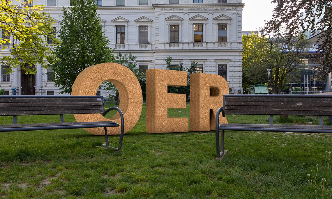 Orange OER-Buchstaben auf einer grünen Wiese mit einer Albau-Fassade eines Unigebäudes im Hintergrund