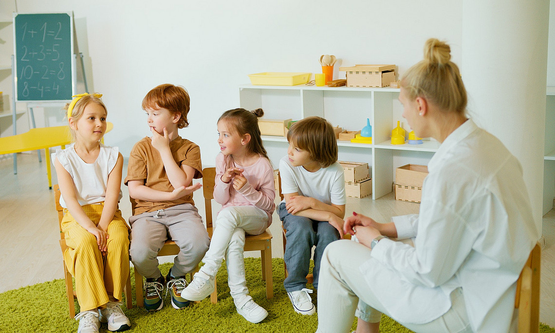 Vier Kinder und eine pädagogische Fachkraft sitzen auf Stühlen in einem Kindergarten.