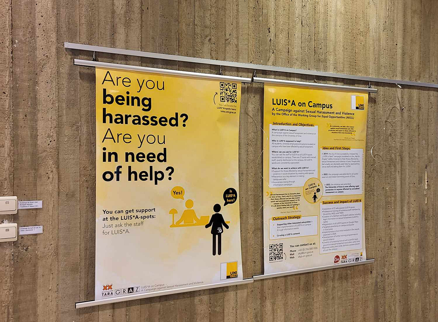 Zwei große Poster hängen an einer braunen Wand, das linke Poster stellt das LUIS*A-Sujet auf Englisch dar, während das rechte Poster auf LUIS*A am Campus als Projekt eingeht ©Yellow White, Belgium