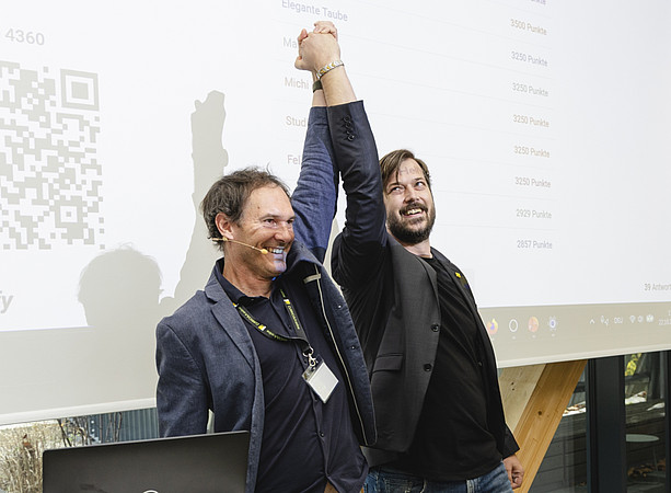 Zwei lächelnde Männer heben gemeinsam die Arme und feiern vor einer Projektionsfolie mit Rangliste und Punktzahlen. ©@foto-gasser.at