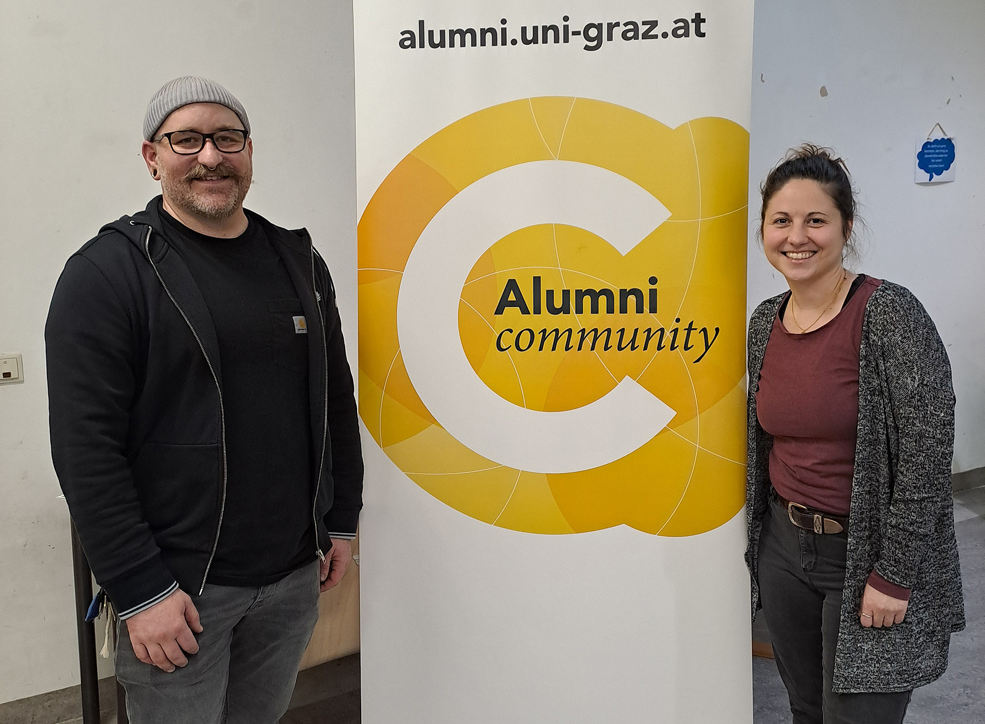 Zwei Personen (Susanne Alter und Stefan Schatz) stehen rechts und links vor einem Roll-up der Alumni Community ©Uni Graz/Verena Marchner