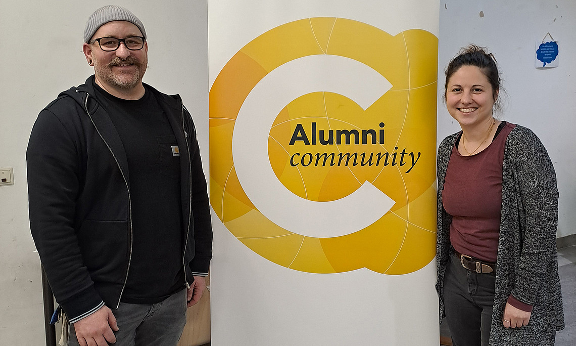 Zwei Personen (Susanne Alter und Stefan Schatz) stehen rechts und links vor einem Roll-up der Alumni Community
