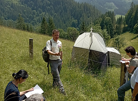 Ein Foto von einer Gruppe von Menschen in den österreichischen Alpen. Wissenschaftler:innen und Parkmitarbeiter:innen treffen sich, um Forschungspläne für wissenschaftliche Geräte zu besprechen, die bei der Feldarbeit verwendet werden sollen Das Foto zeigt grasbewachsene Hügel mit einigen Bäumen im Hintergrund. Ein Mann steht auf und hält Alpenblumen 