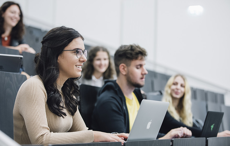 Eine Studierende sitzt mit StudienkollegInnen und Laptop im Hörsaal und hört lächelnd dem Vortrag zu.