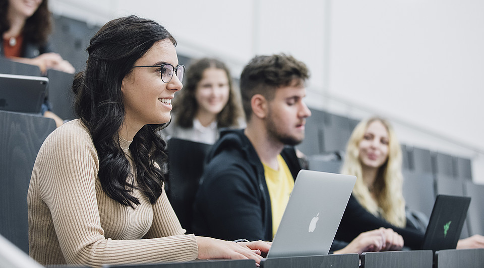 Eine Studierende sitzt mit StudienkollegInnen und Laptop im Hörsaal und hört lächelnd dem Vortrag zu.