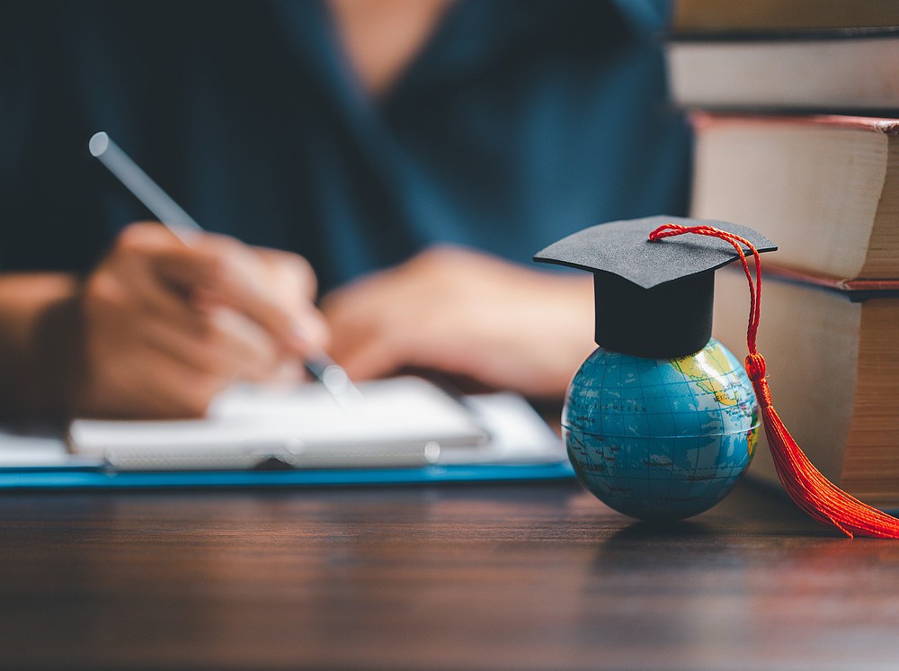 close-up of a graduate signing a form in front a small globe with a stack of books ©By JD8; stock.adobe.com 