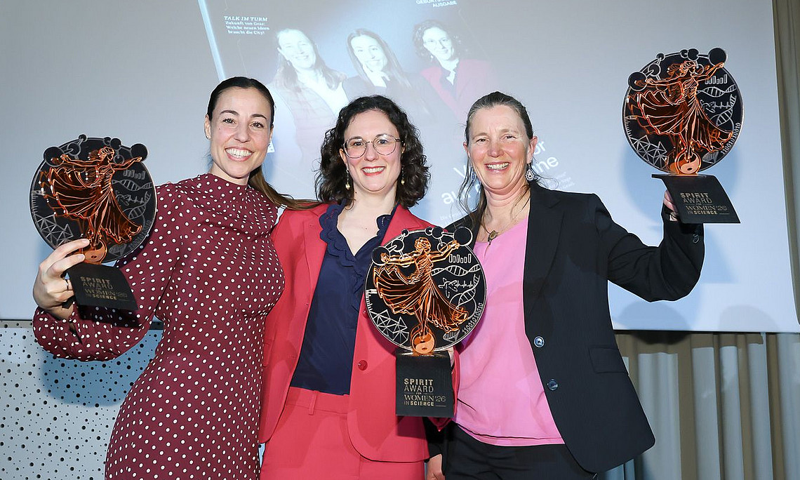 Three people stand side by side in festive clothing, holding large, round trophies up to the camera. Each trophy shows a stylized, dancing figure in copper color against a dark background. The people are wearing suits and a polka-dot dress; the clothing is in shades of red, blue, and black. In the background, there is a screen with the word “spirit” and a magazine or presentation graphic. The scene looks like an award ceremony in an indoor setting.