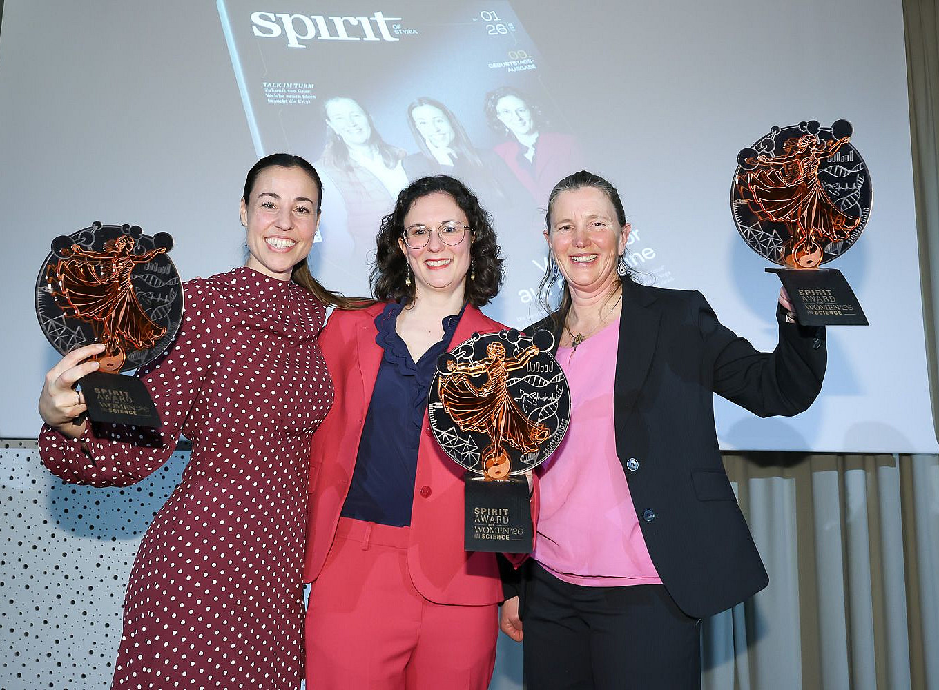 Drei Personen stehen nebeneinander in festlicher Kleidung und halten große, runde Trophäen in die Kamera. Jede Trophäe zeigt eine stilisierte, tanzende Figur in Kupferfarbe vor dunklem Hintergrund. Die Personen tragen Anzüge und ein gepunktetes Kleid; die Kleidung ist in Rot-, Blau- und Schwarztönen gehalten. Im Hintergrund ist eine Leinwand mit dem Wort „spirit“ und einer Magazin- oder Präsentationsgrafik zu sehen. Die Szene wirkt wie eine Preisverleihung in einem Innenraum. 