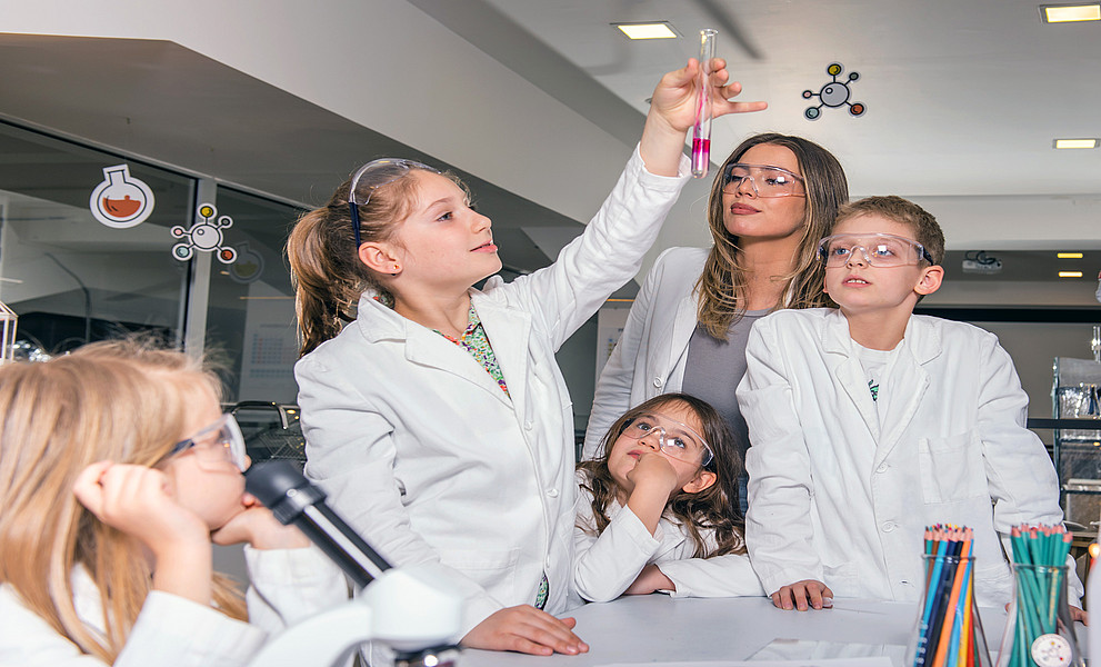 drei Mädchen, ein Bub, eine junge Lehrerin in weißen Arbeitsmänteln; ein Mädchen hält ein Reagenzglas mit einer roten Flüssigkeit hoch