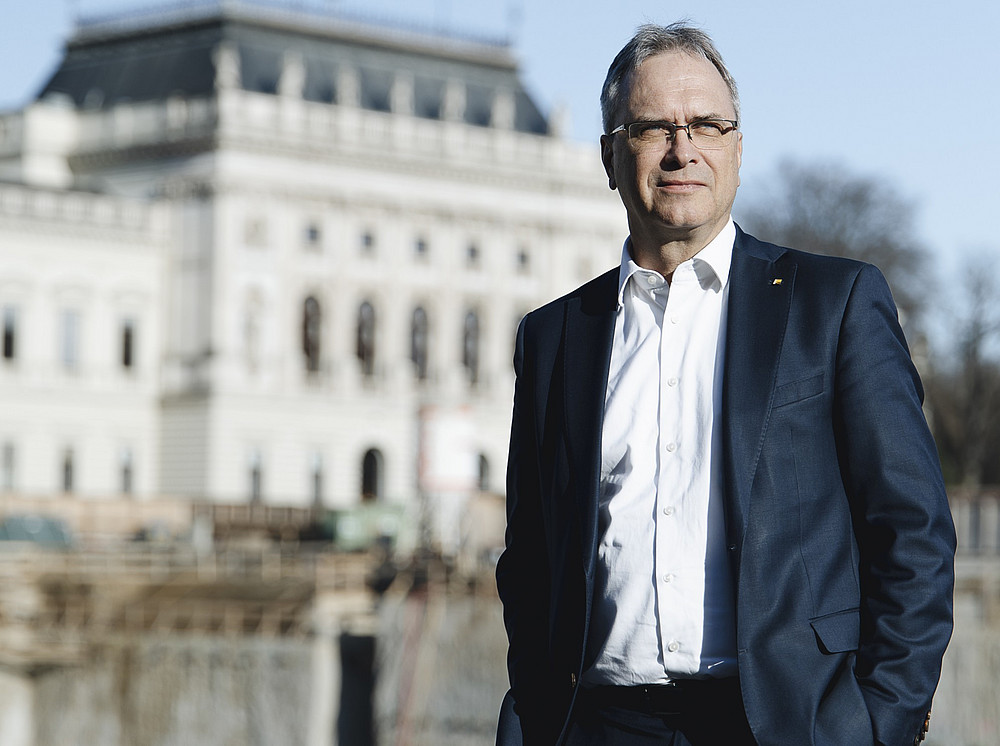 Mann in dunklem Anzug und weißem Hemd steht vor historischer Gebäudefassade, im Vordergrund Baustelle unter blauem Himmel. 