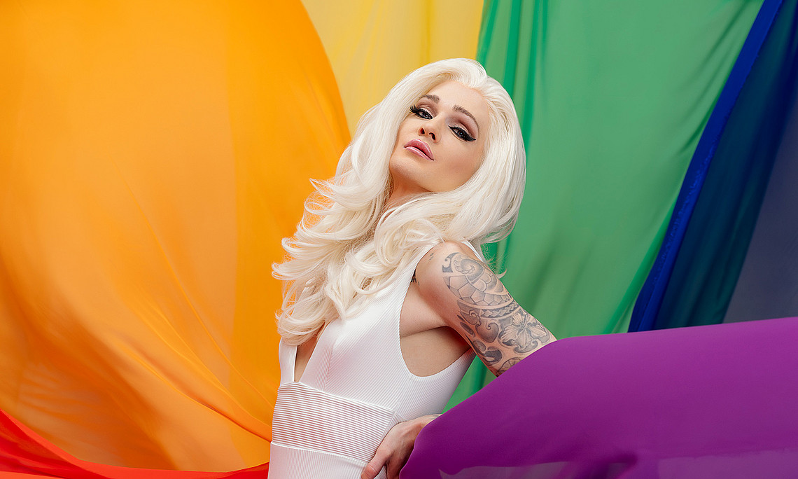 Drag Queen Gloria Hole mit weißem Kleid in regenbogenfarbigen Tüchern