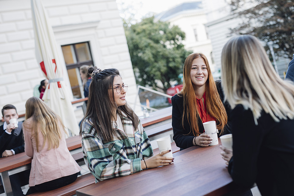 Studierende sitzen mit To-Go-Heißgetränken gemeinsam im Außenbereich eines Uni-Cafés an einem Tisch und unterhalten sich fröhlich.