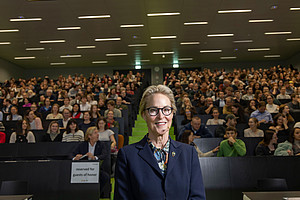 A woman in a business suit stands in front of a large lecture hall with modern lighting, filled with a large number of people.