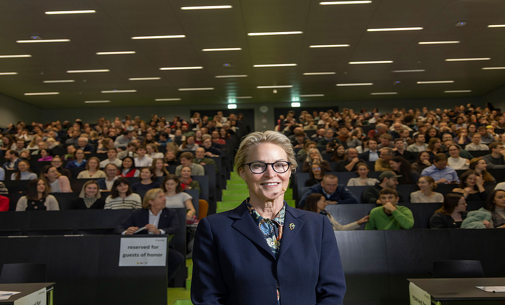 Eine Frau in einem Business-Kostüm steht vor einem großen Hörsaal mit moderner Beleuchtung, gefüllt mit einer Vielzahl von Menschen.