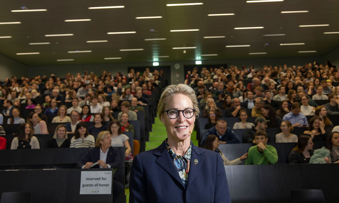 A woman in a business suit stands in front of a large lecture hall with modern lighting, filled with a large number of people.
