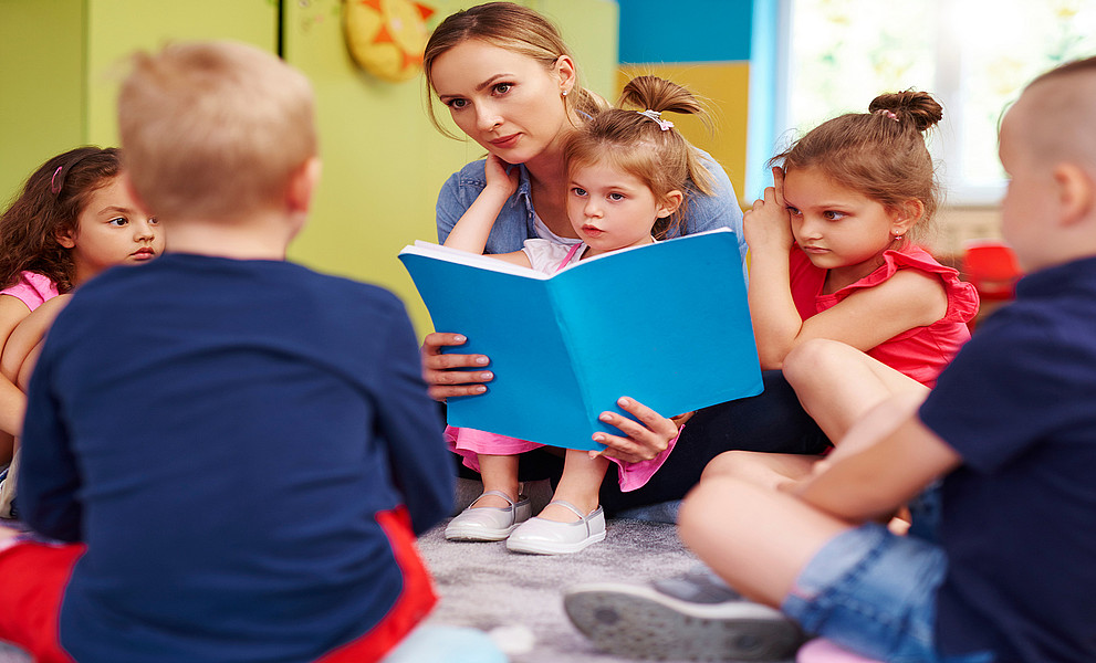 Kindergartenkinder sitzen im Kreis am Boden, eine Frau liest aus einem Buch vor