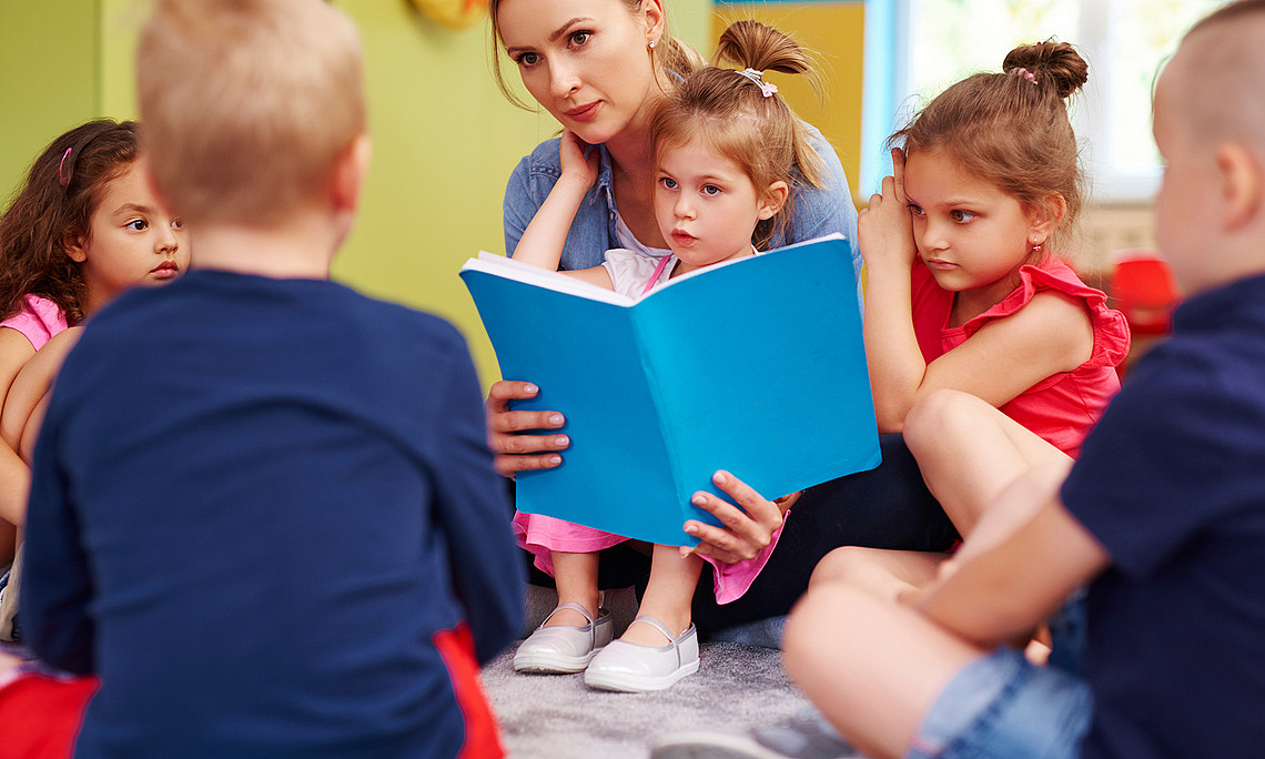 Kindergarten children sit in a circle on the floor, a woman reads from a book