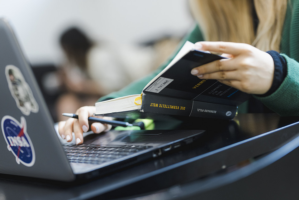 Studierende liest im Buch "Die vermittelte Welt" und arbeitet an ihrem Laptop