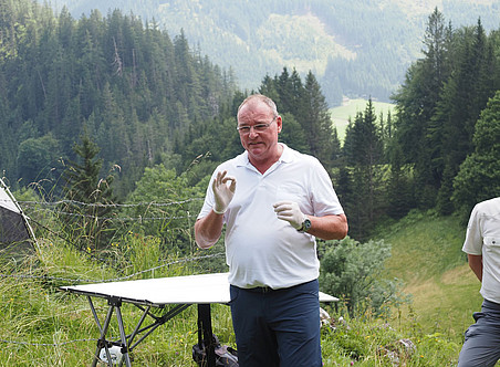 Foto eines Mannes in den Fünfzigern, der ein weißes Polohemd und eine blaue Hose trägt und auf dem Gipfel eines Berges eine Präsentation hält. Er steht an einem Tisch mit einem kleinen schwarzen Bildschirm und zeigt Handgesten, während er mit einer Gruppe von Leuten hinter ihm spricht. ©Nationalpark Gesäuse/Wölger